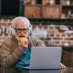 senior man with headphones on, eating apple, talking to someone using his laptop