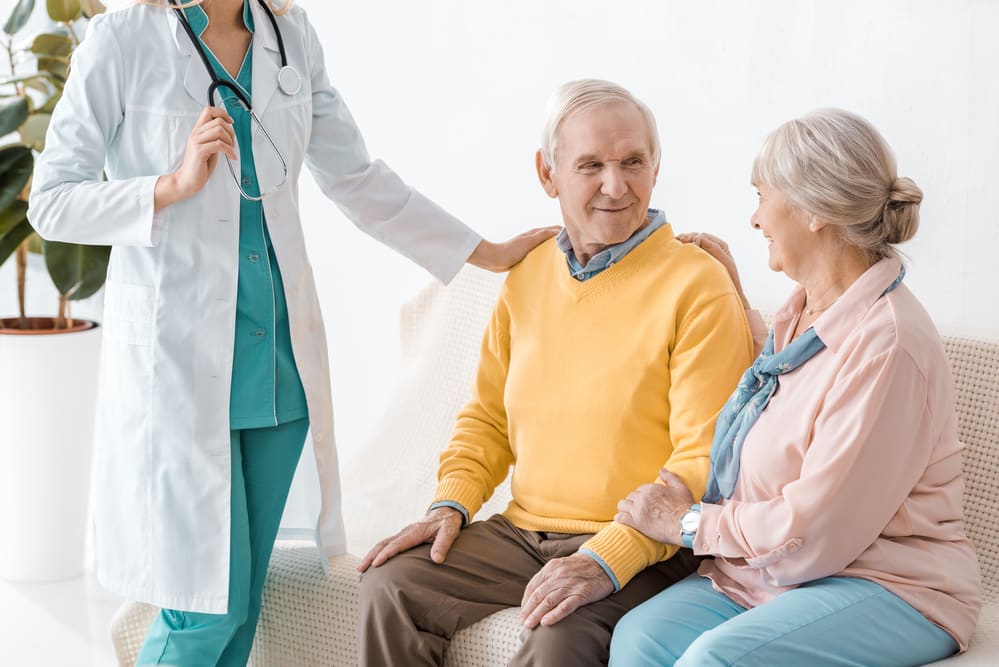 Smiling senior couple sitting on couch, young nurse with hand on man's shoulder