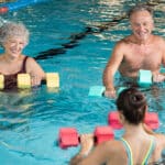 Two seniors taking water aerobics class from young instructor in indoor pool