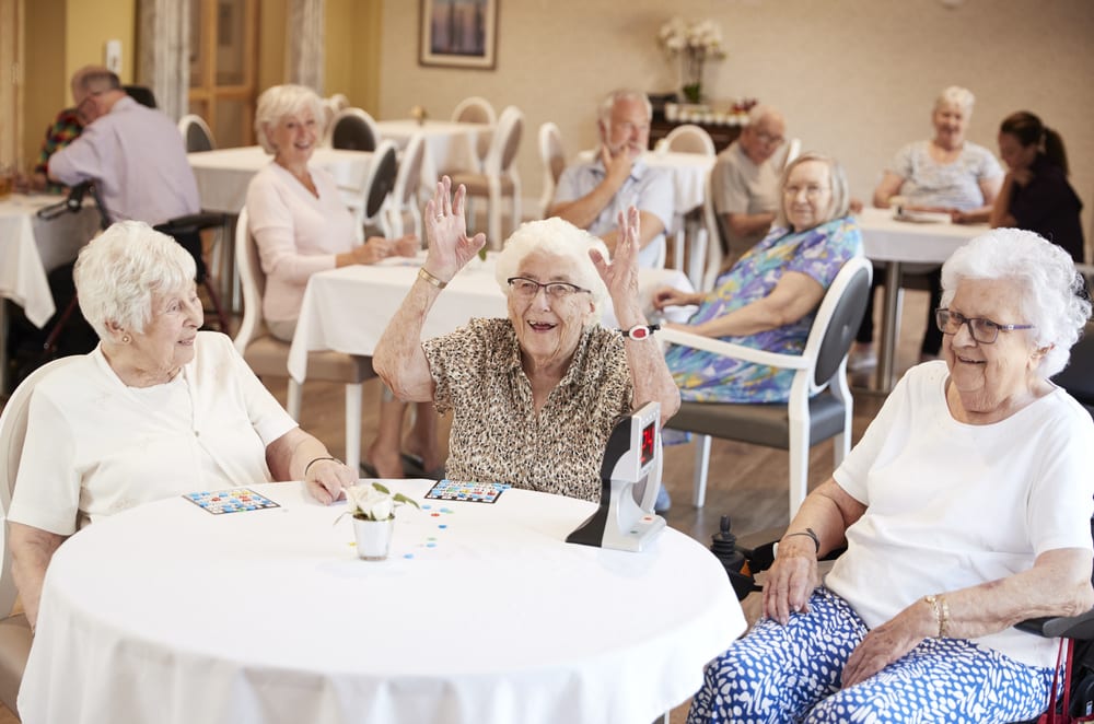 Three senior women at table playing bingo, one with hands in the air, other people in background